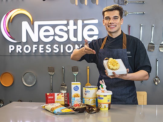 Chef Pedro Liberalesso posa junto a uma bancada segurando uma embalagem com ovo de Páscoa recheado, preparado com os produtos
