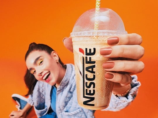 Mulher de cabelo preso e vestindo blusa azul sorri enquanto segura bebida de nescafé gelado; fundo da imagem é totalmente lar