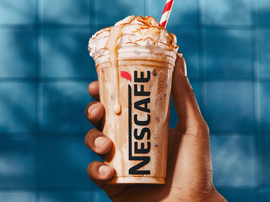 Copo de nescafé gelado transborda; a bebida tem tons de bege e branco, e o copo é segurado apenas por uma mão; fundo de azule