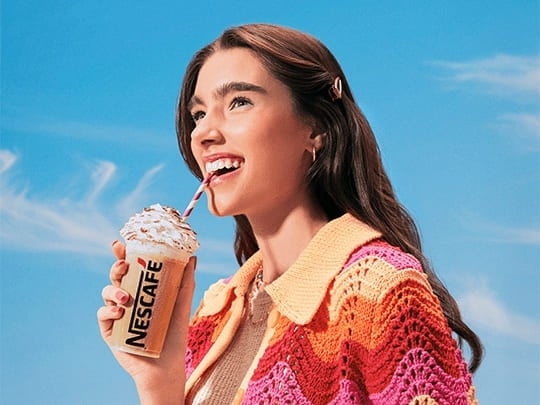 Mulher bebe de canudo bebida nescafé gelado; ela veste casaco de tricot com cores quentes; ao fundo, um céu azul