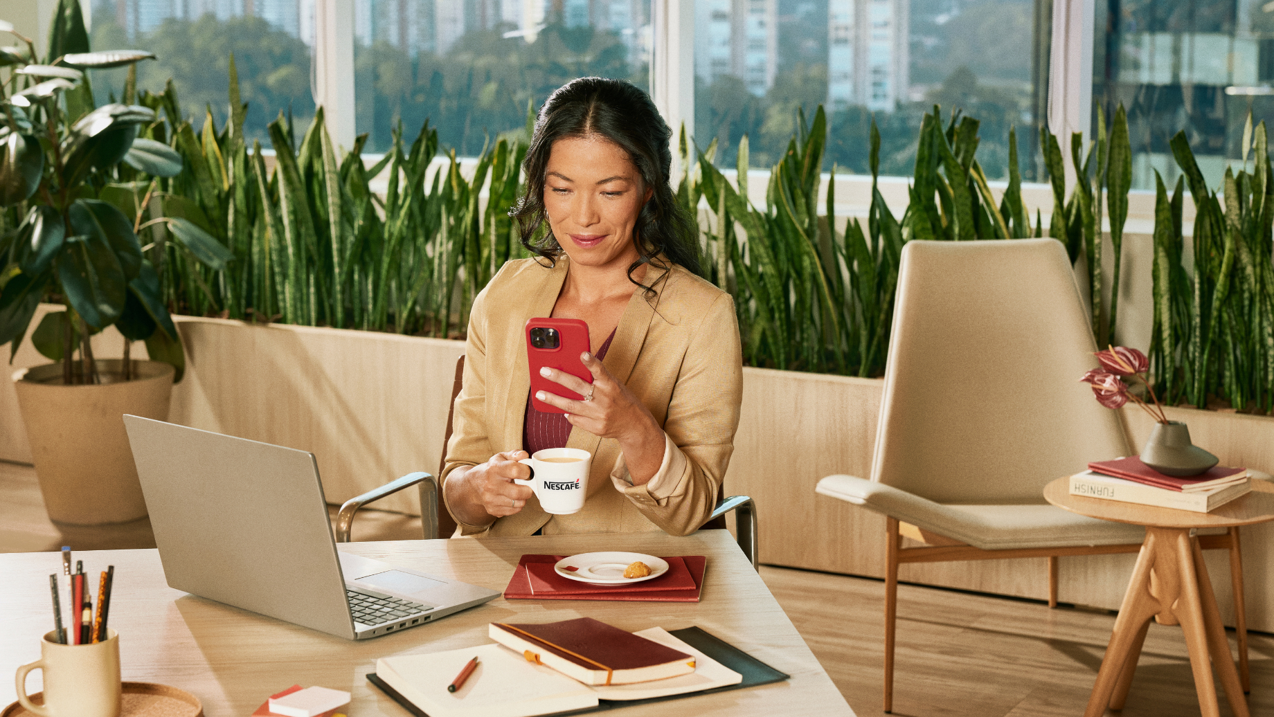 Pessoa sentada em mesa de escritório usando celular e segurando xícara de café Nescafé, com notebook, caderno e plantas ao fu