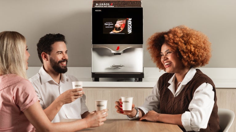 duas mulheres e um homem sentados sorrindo em torno de uma mesa, segurando copos de café expresso de uma máquina de café