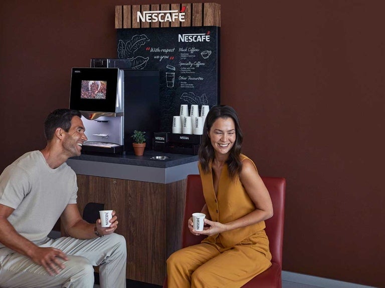 Homem e mulher se sentam no cantinho do café da empresa, que tem uma máquina Nescafé, e sorriem