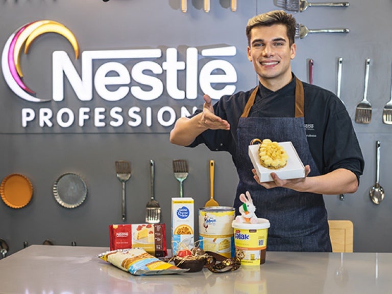 Chef Pedro Liberalesso posa junto a uma bancada segurando uma embalagem com ovo de Páscoa recheado, preparado com os produtos