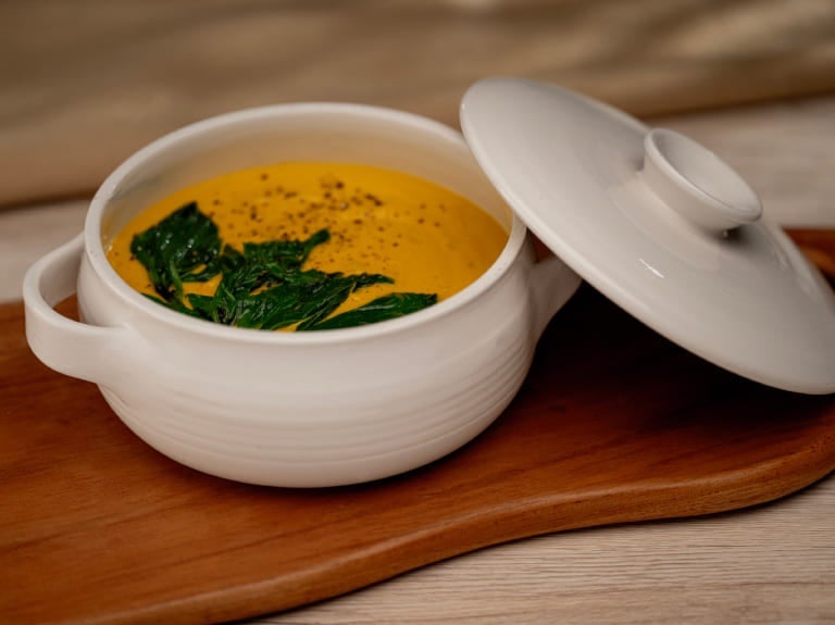 sopa de abóbora servida em uma tigela de cerâmica branca sobre uma tábua de madeira.