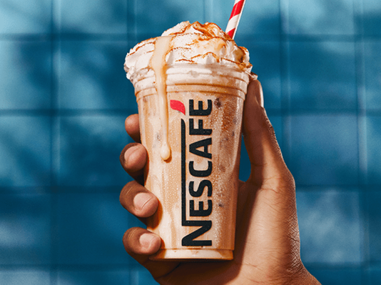 Copo de nescafé gelado transborda; a bebida tem tons de bege e branco, e o copo é segurado apenas por uma mão; fundo de azule