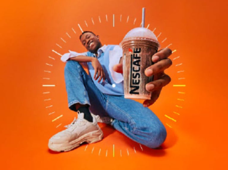 homem agachado, de calça jeans e blusa azul, segura copo com bebida nescafé gelado e aponta para a foto; a imagem tem fundo l