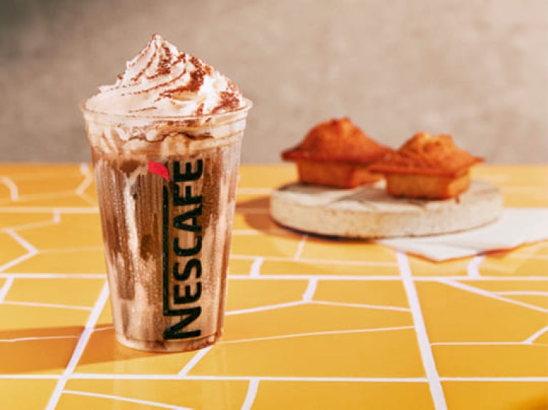 Bebida de nescafé gelado sobre uma mesa; ao fundo, há dois bolinhosque acompanham a bebida