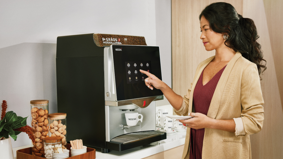 Pessoa utilizando máquina de café Nescafé em escritório, tocando painel digital para preparar bebida; xícaras e acompanhament