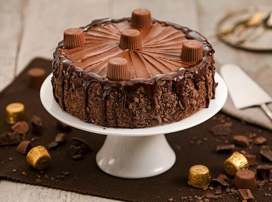 Bolo de chocolate coberto com bombons Alpino, está sob um prato branco, apoiado em uma mesa que tem mais bombons Alpino espal