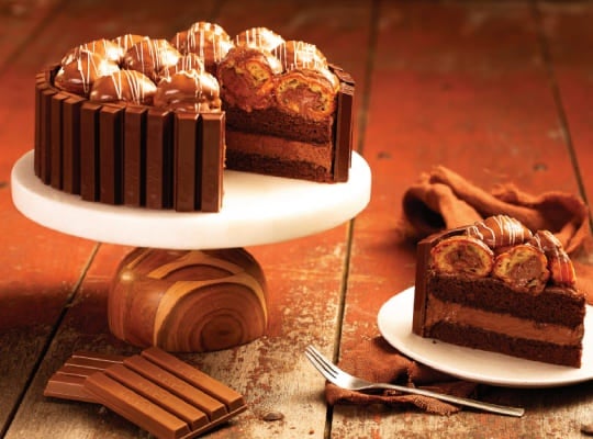 Alt text: Imagem de bolo de carolina feito com KitKat. O bolo está em um suporte, e teve uma fatia cortada.