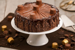 Bolo de chocolate coberto com bombons Alpino, está sob um prato branco, apoiado em uma mesa que tem mais bombons Alpino espal