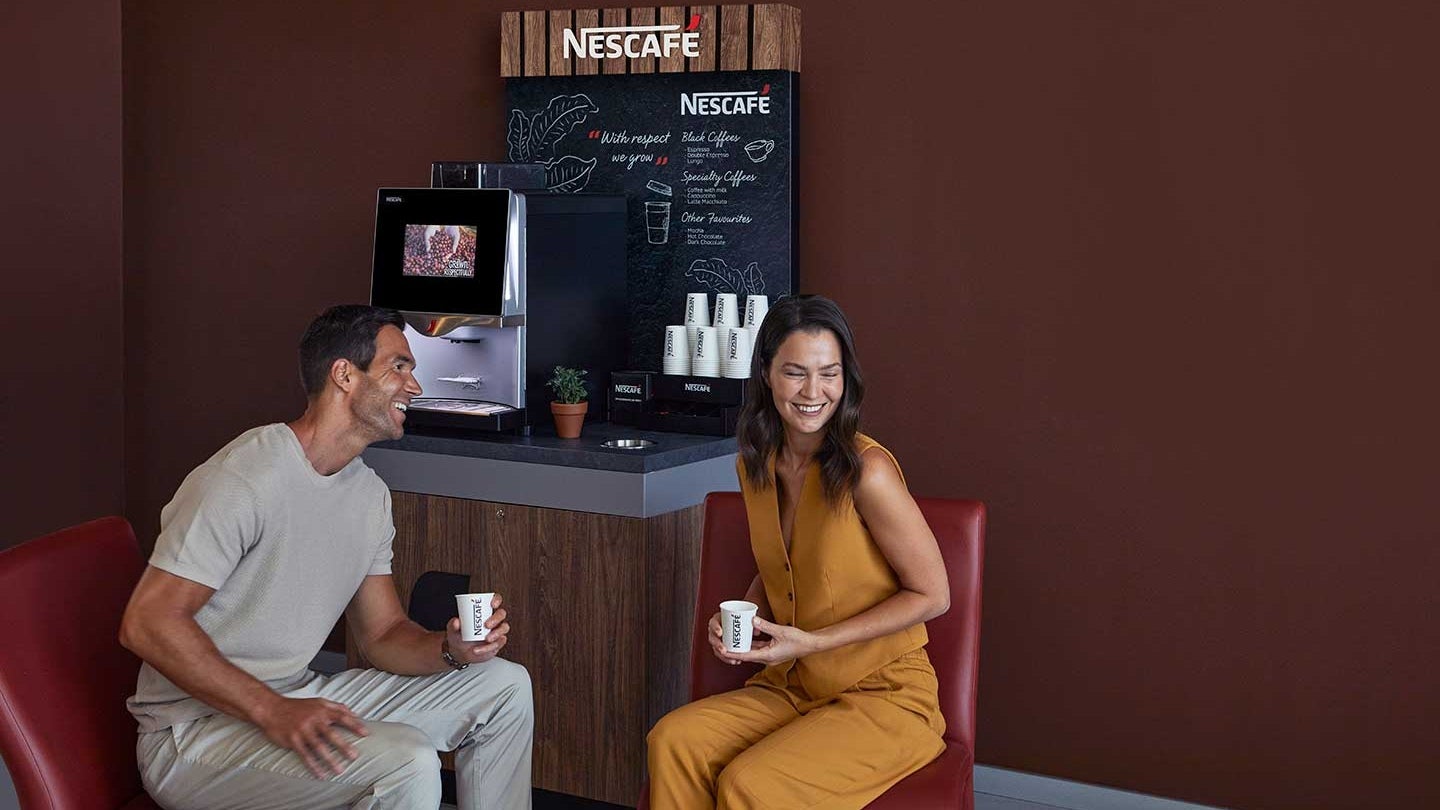Homem e mulher se sentam no cantinho do café da empresa, que tem uma máquina Nescafé, e sorriem