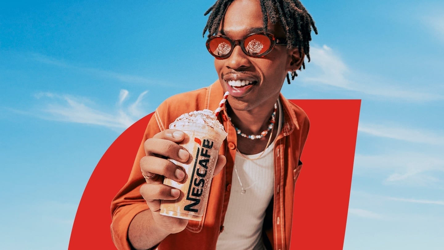 Homem negro de óculos escuros de lentes vermelhas toma bebida nescafé gelado; ele veste uma camisa vermelha por cima de uma r